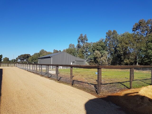 A wide shot of a fenced paddock area with a modern grey barn, surrounded by trees and a clear blue sky.