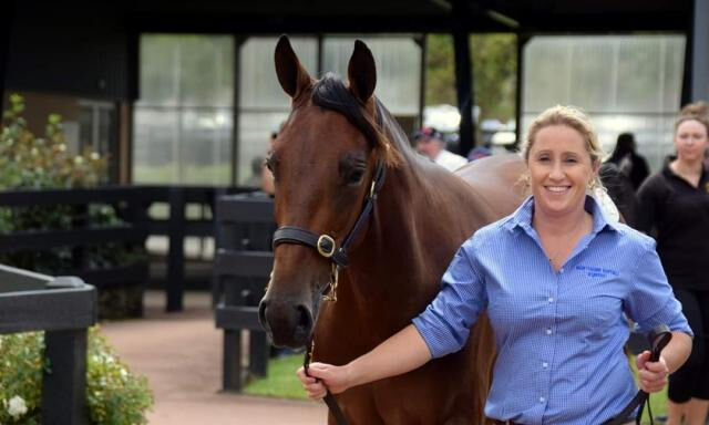 A woman in a blue shirt smiling while leading a brown horse by a halter in an outdoor area, with other people in the background.